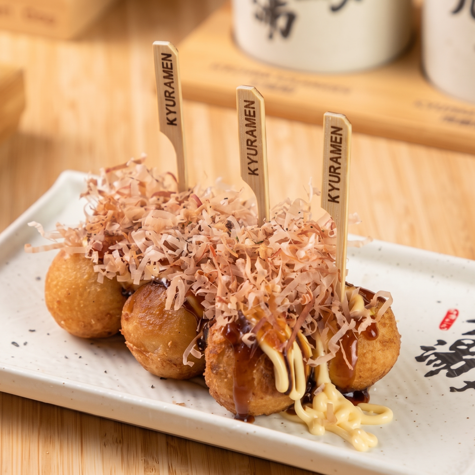 Takoyaki balls topped with bonito flakes, sauces, and Kyoramen-branded picks on a Japanese ceramic plate.