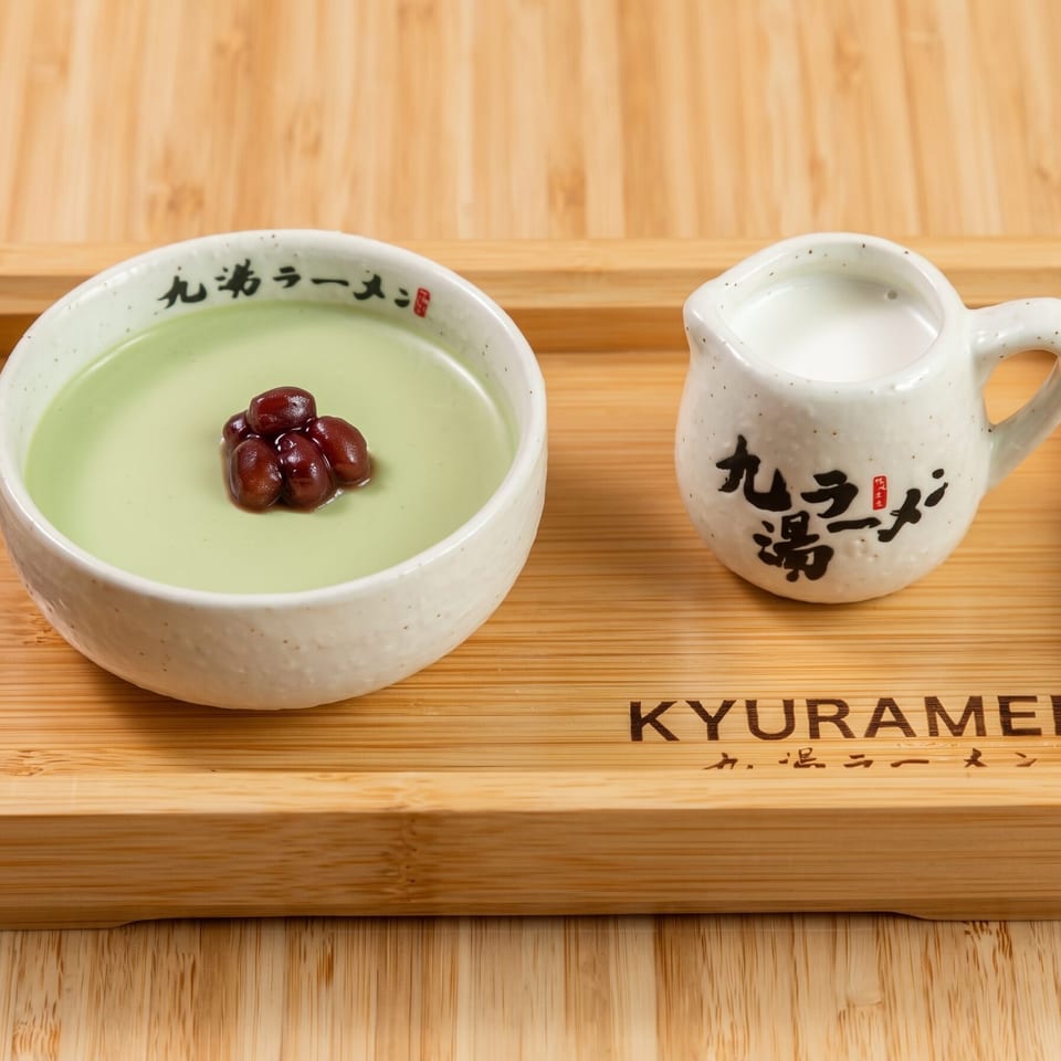 Pale green matcha dessert topped with red beans beside a ceramic pitcher on a wooden Kyuramen tray.