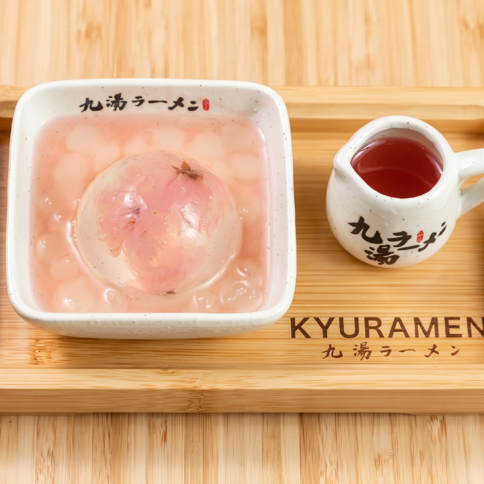 Kyuramen-branded dessert bowl with a pink flower-shaped confection and red sauce pitcher on a wooden tray.