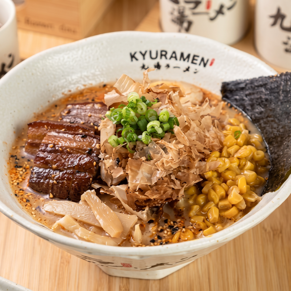 Kyuramen ramen bowl topped with glazed pork, corn, green onions, bamboo shoots, seaweed, and bonito flakes.